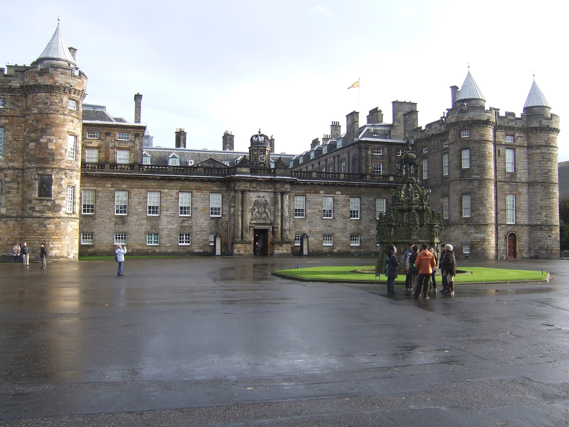 Visitors cross the wet forecourt of Edinburgh's Palace of Holyroodhouse, with its round towers and ornate fountain.