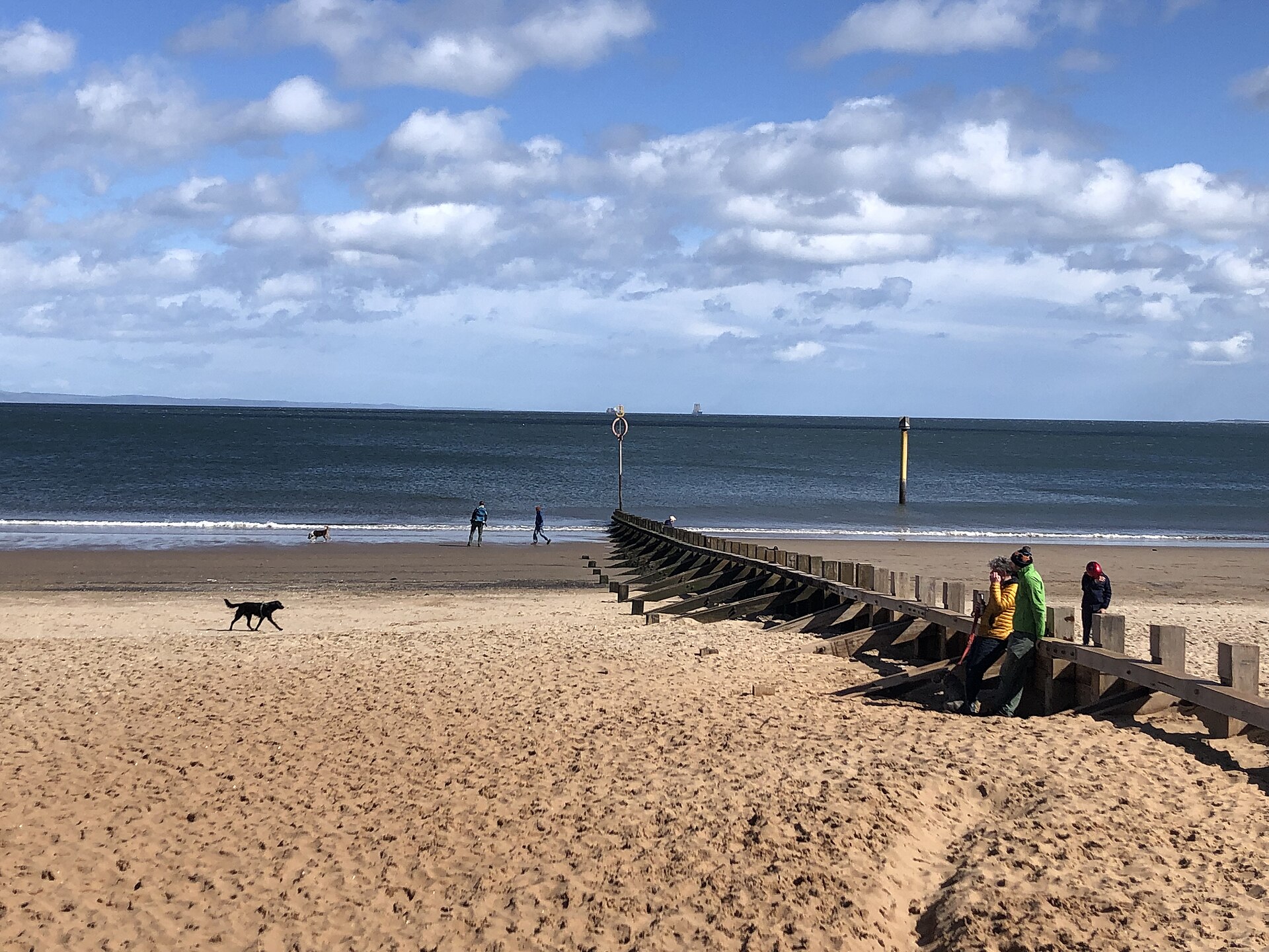 Visitors and dogs cross Portobello Beach beside a timber groyne as small waves reach the Edinburgh shoreline.