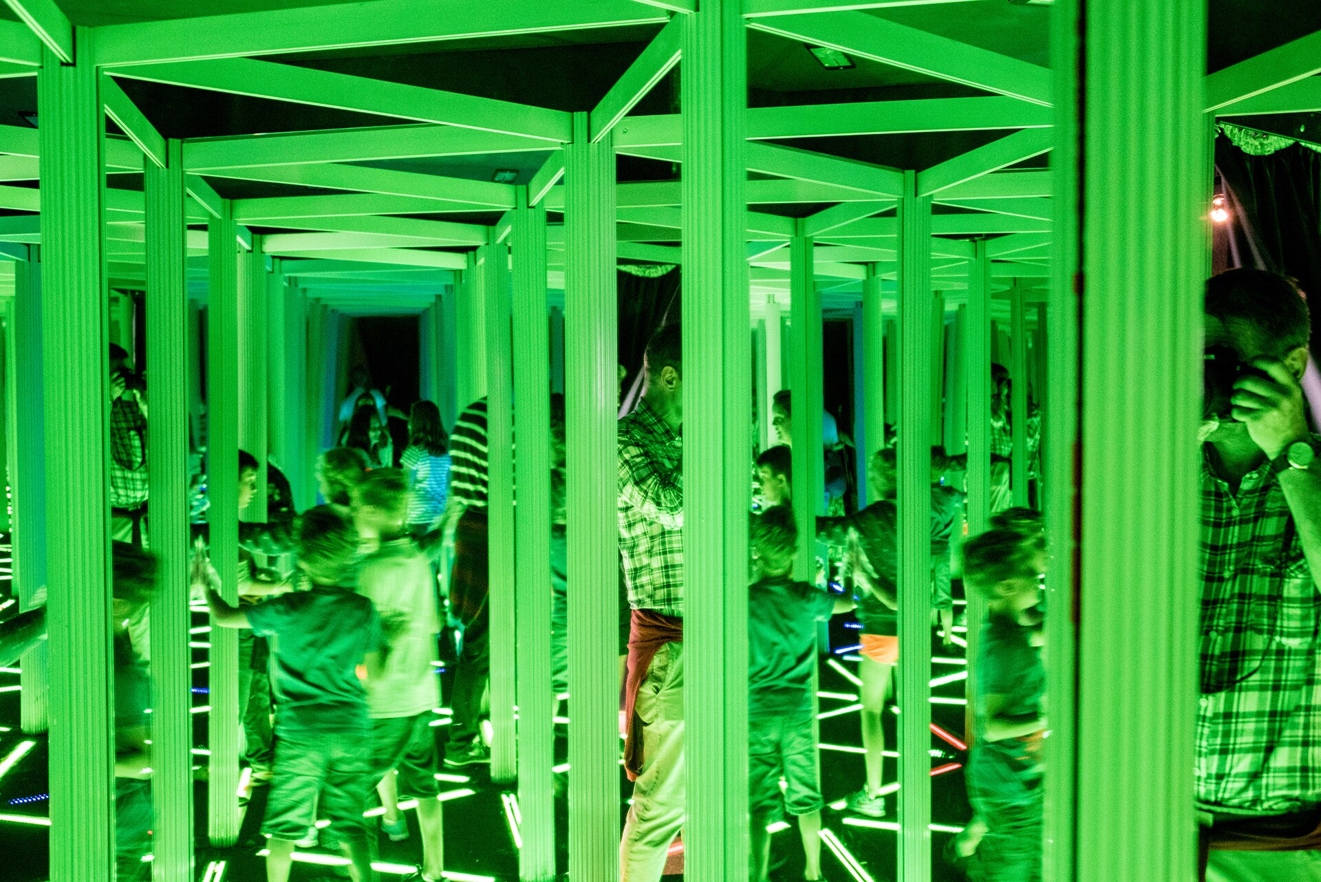 Visitors explore a green-lit mirror maze at Camera Obscura in Edinburgh, reflected repeatedly between rows of rectangular frames.