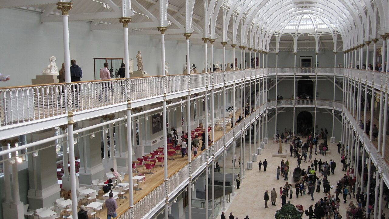 Visitors explore the National Museum of Scotland beneath white iron balconies and the high glazed roof of its central hall.