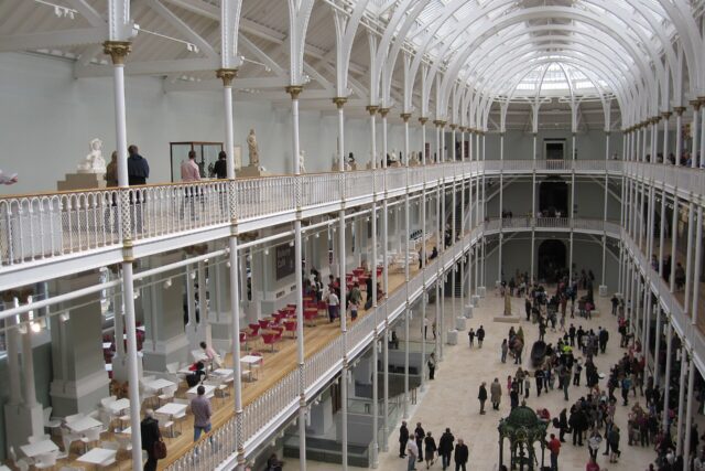 Visitors explore the National Museum of Scotland beneath white iron balconies and the high glazed roof of its central hall.