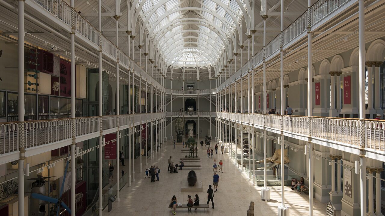 Visitors explore the National Museum of Scotland Grand Gallery beneath a vaulted glass roof and white iron balconies.