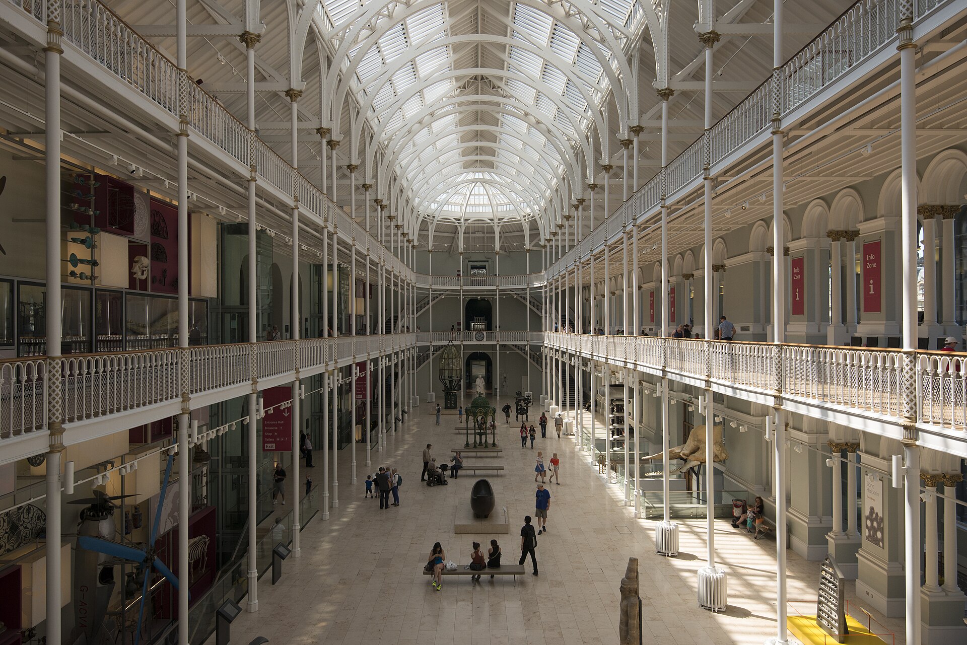 Visitors explore the National Museum of Scotland Grand Gallery beneath a vaulted glass roof and white iron balconies.