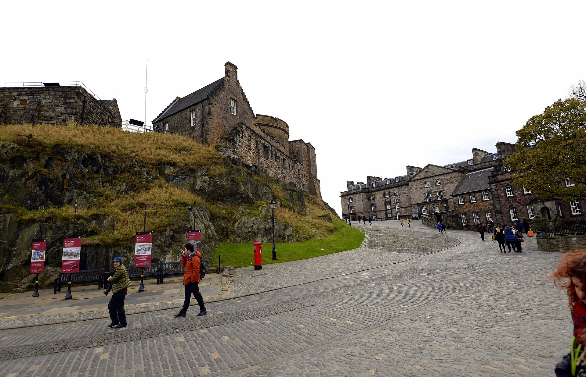 Visitors follow a cobbled path uphill through Edinburgh Castle beside a rocky bank and stone buildings.