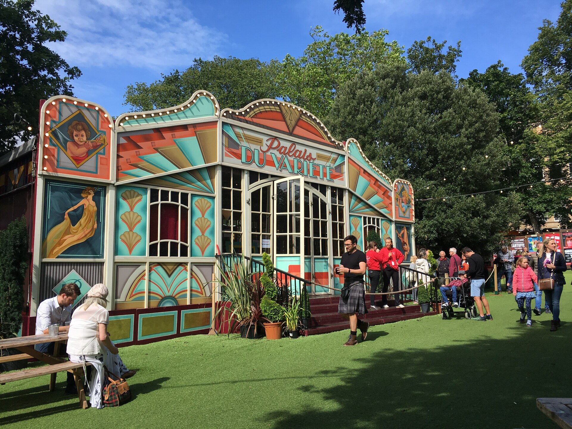 Visitors gather outside a colourful Spiegeltent with ornate painted panels and glazed doors in Edinburgh's leafy George Square Gardens.