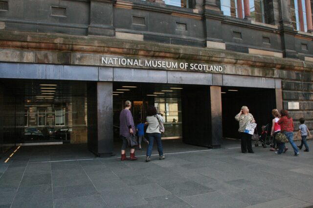 Visitors gather outside the National Museum of Scotland entrance on Chambers Street beneath lettering naming the museum.