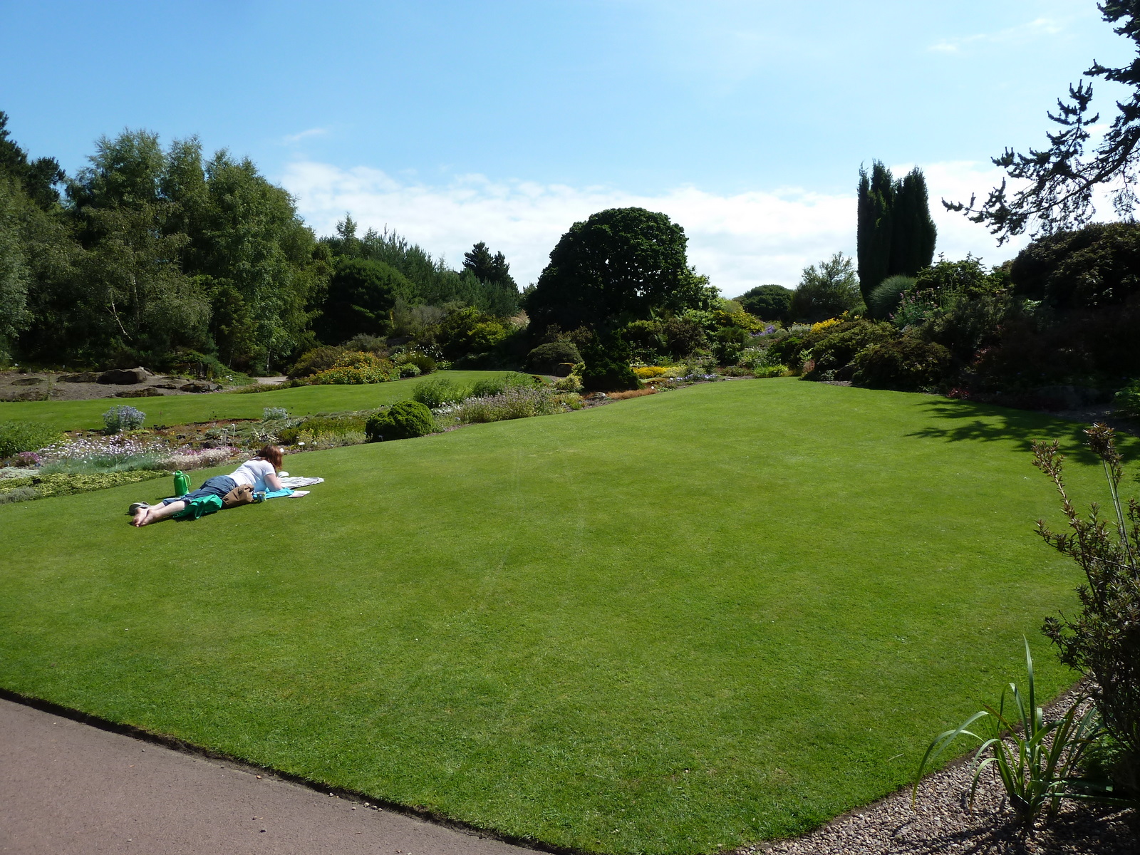 Visitors relax on a broad green lawn beside flower beds and mature trees at the Royal Botanic Garden Edinburgh.