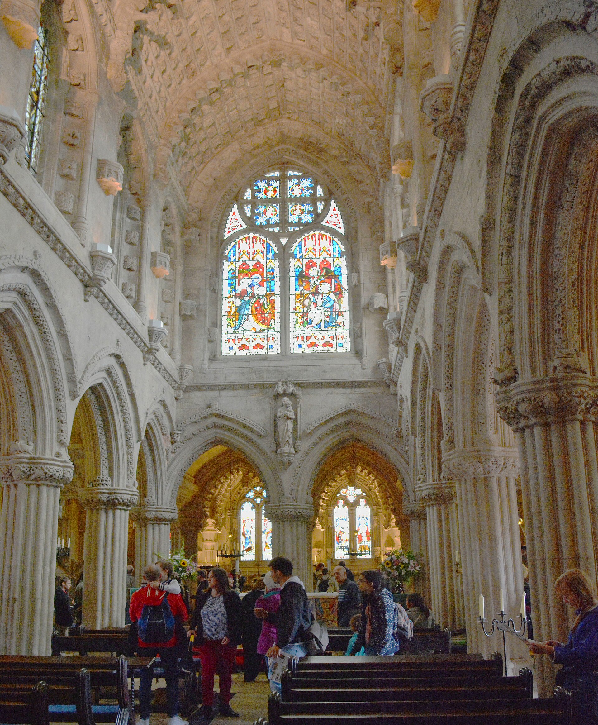 Visitors stand beneath carved sandstone arches and vaulting inside Rosslyn Chapel, with stained glass above the pews.