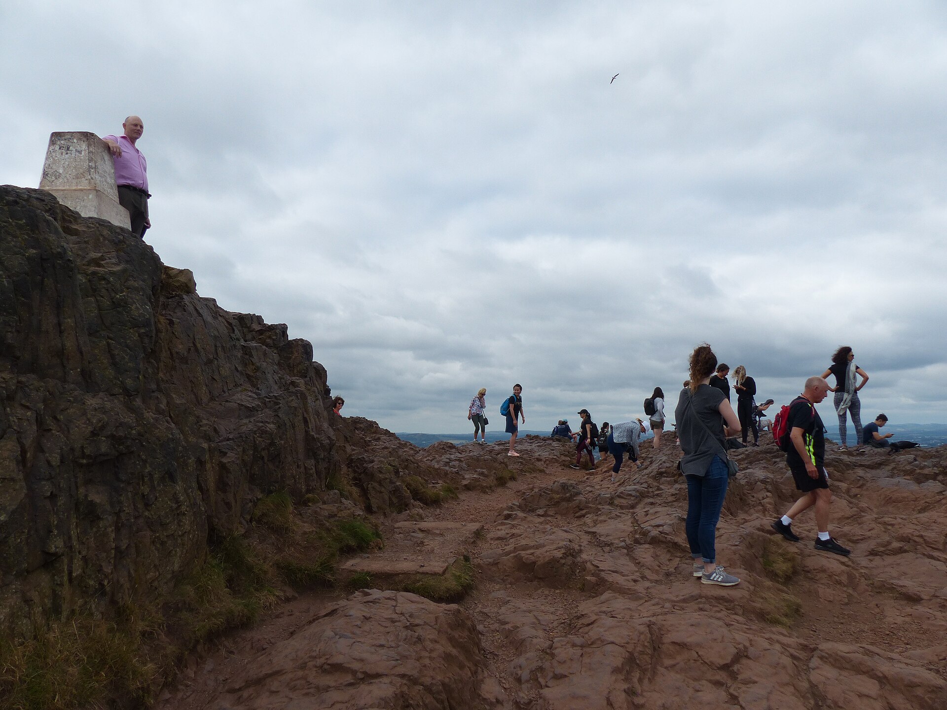 Walkers gather on the rocky summit of Arthur's Seat in Edinburgh beside a stone marker under cloudy skies.