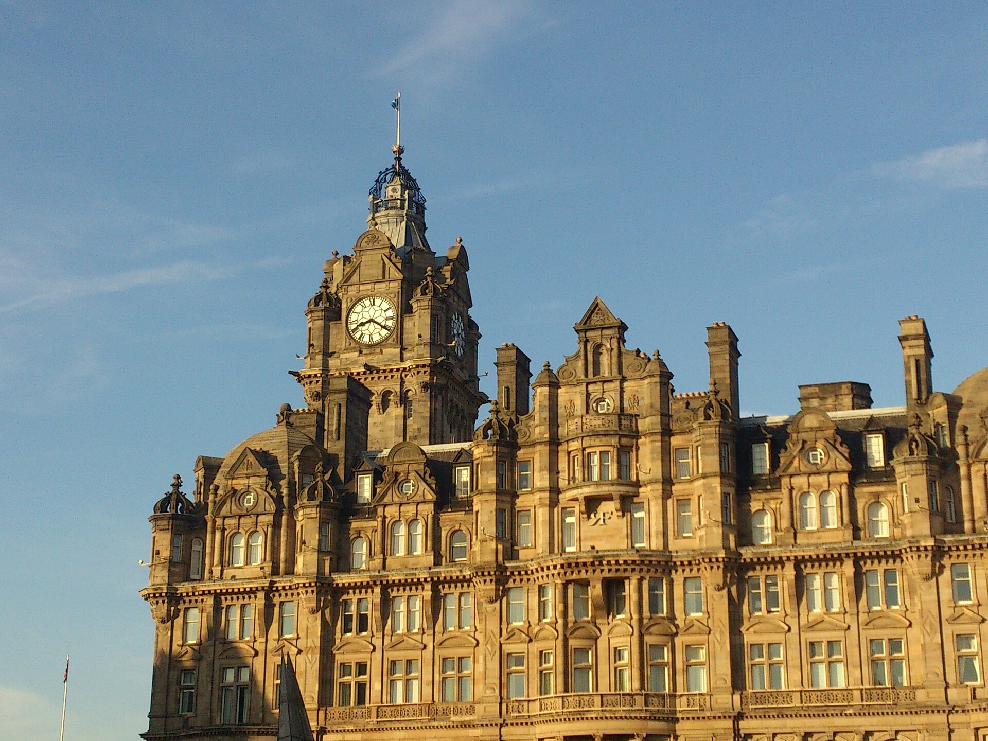 Warm sunlight illuminates the Balmoral Hotel clock tower and ornate sandstone facade in Edinburgh beneath a blue sky.