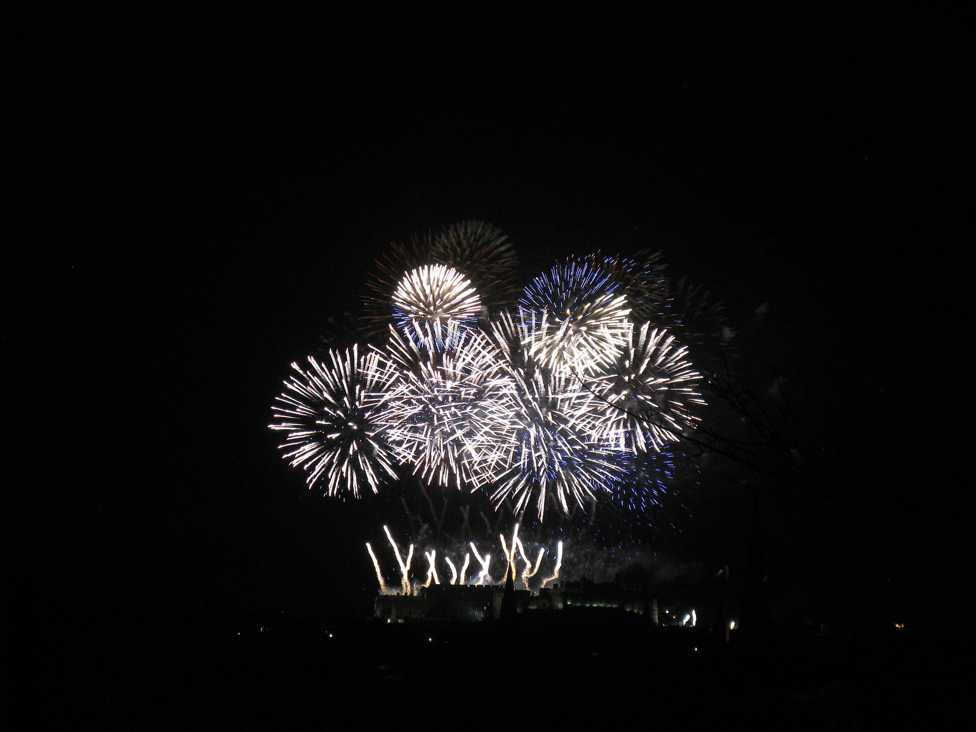 White and blue New Year fireworks spread above Edinburgh Castle, with bright fountains along the castle skyline.